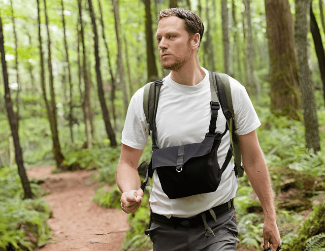 A white man in trekking attire hikes in the woods with a chest bag across his chest, the bag's sleek black fabric contrasting against the man's white hiking shirt.